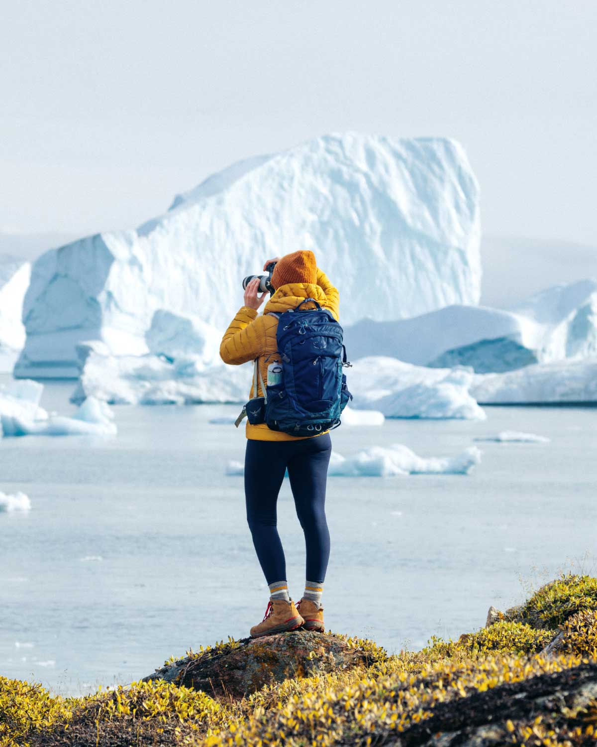 Une personne en tenue de randonnée jaune et bonnet orange prend une photo avec un appareil photo devant un grand iceberg en Arctique ou Antarctique.