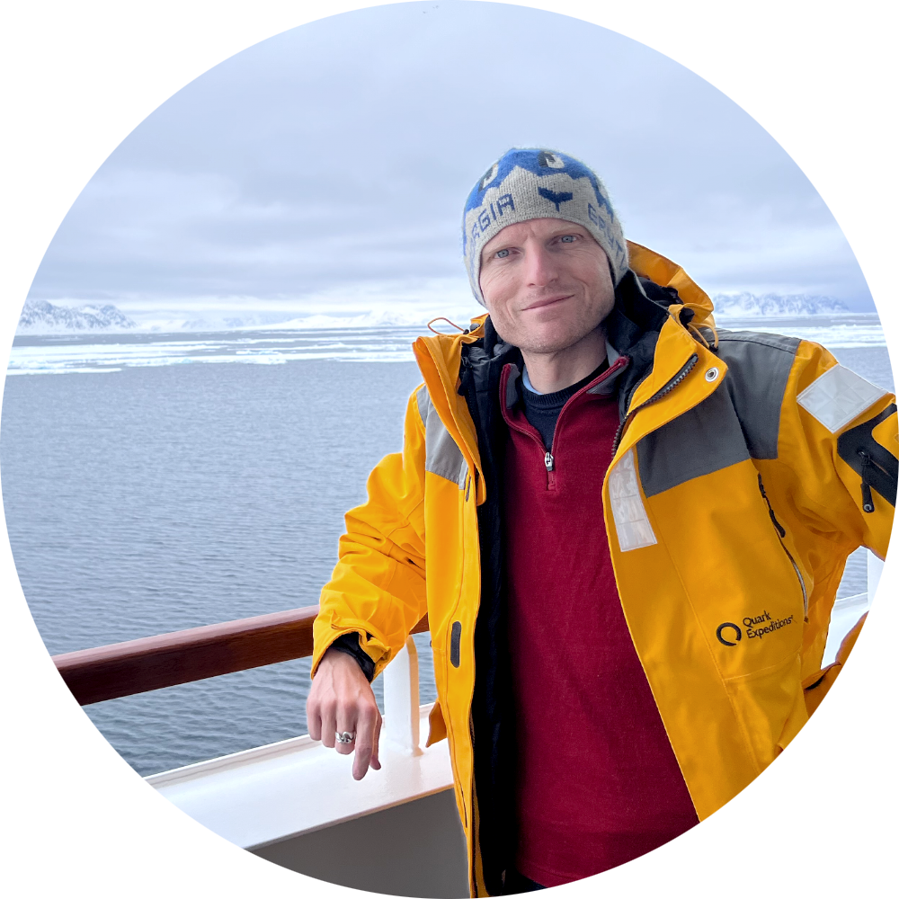 Un homme portant un bonnet bleu et une veste jaune de Quark Expeditions, debout sur le pont d'un bateau avec un paysage arctique en arrière-plan.