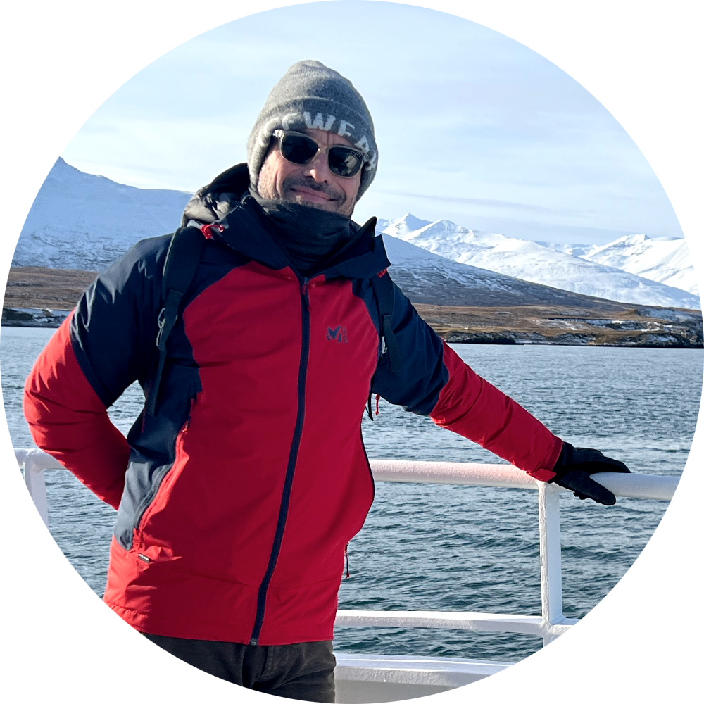 Homme portant un bonnet gris, des lunettes de soleil, une veste rouge et noire, debout sur un bateau avec un paysage de montagnes enneigées en arrière-plan.