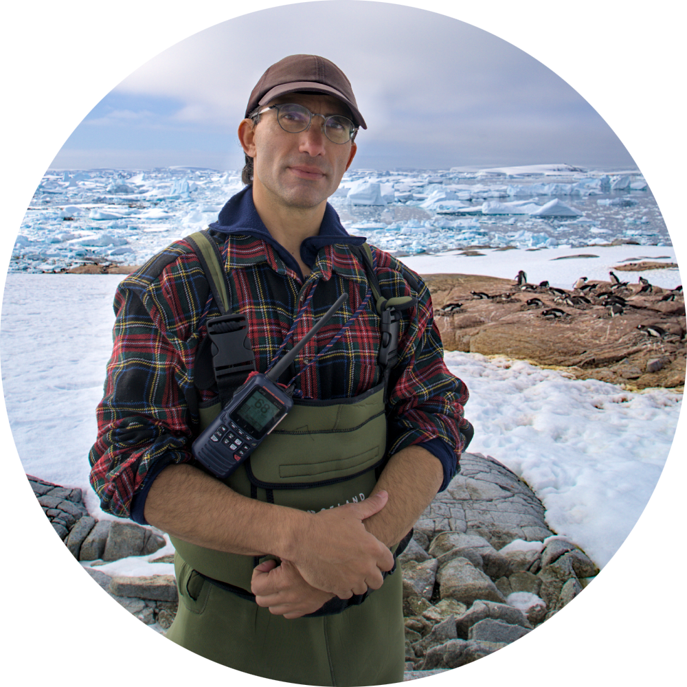 Un homme portant des lunettes et une casquette, vêtu d’une chemise à carreaux et d’une salopette verte avec un talkie-walkie, debout sur des rochers enneigés près de la mer glacée avec des manchots en arrière-plan.